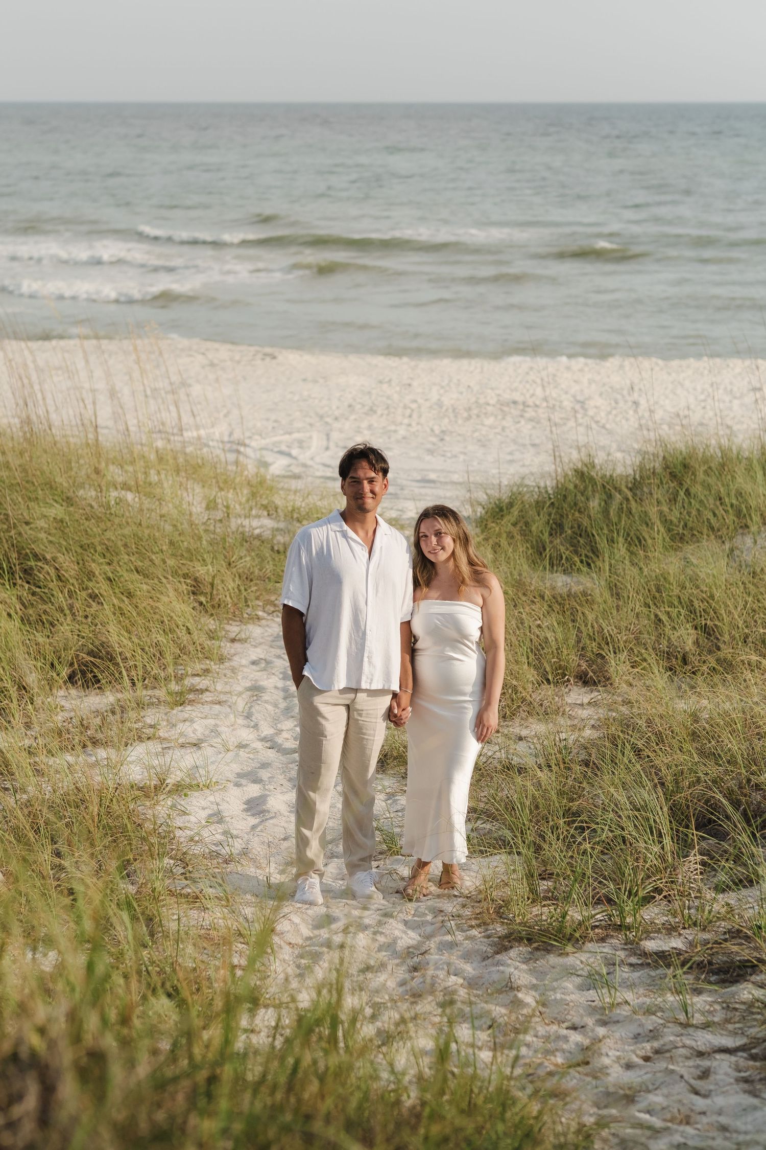 Couple in white and beige outfits standing on beach path with sea grass.
