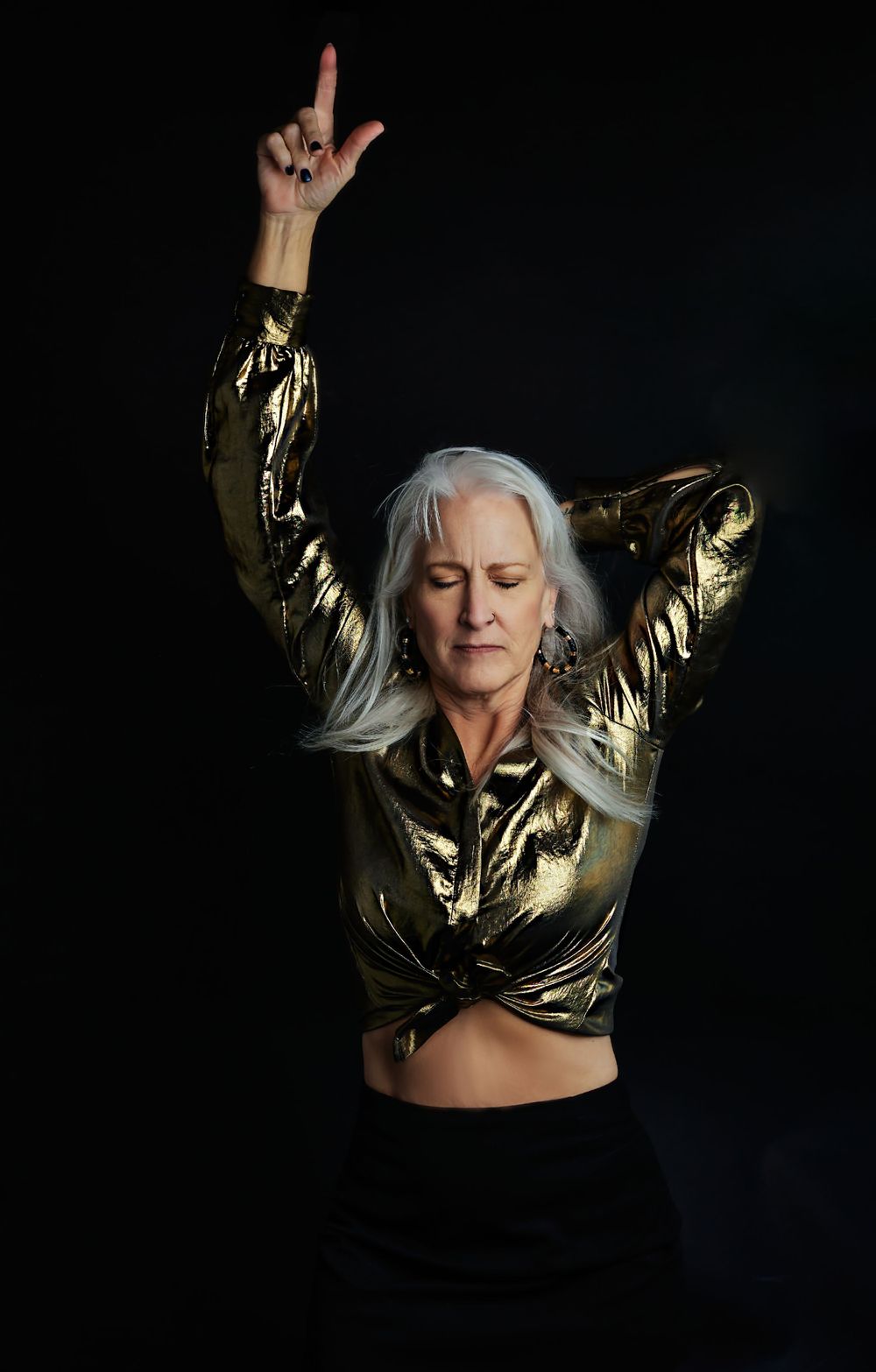 Dynamic pose of figure in metallic gold crop top with arms raised against black background.