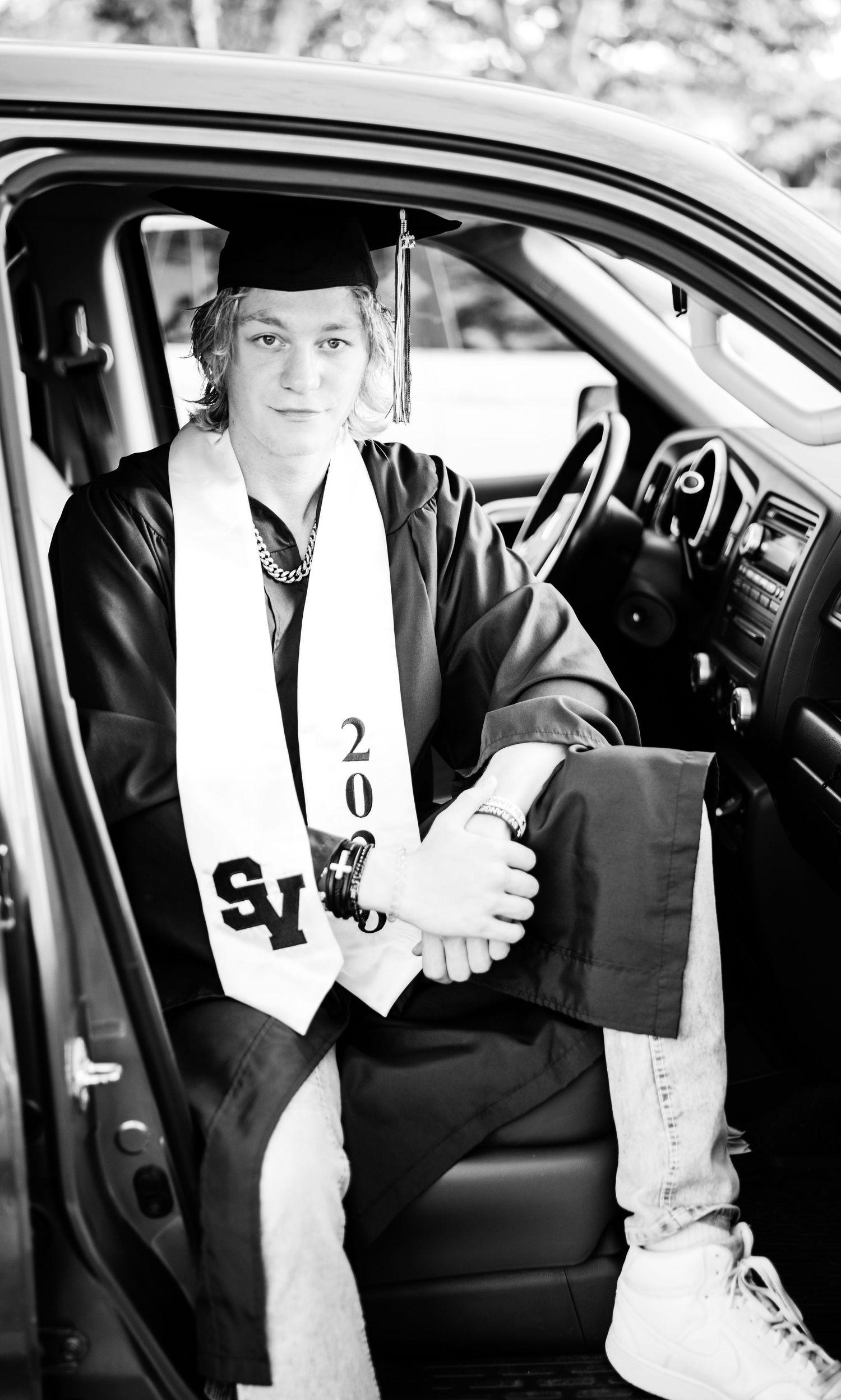 Black and white portrait of a male graduate in cap and gown sitting in a truck door.