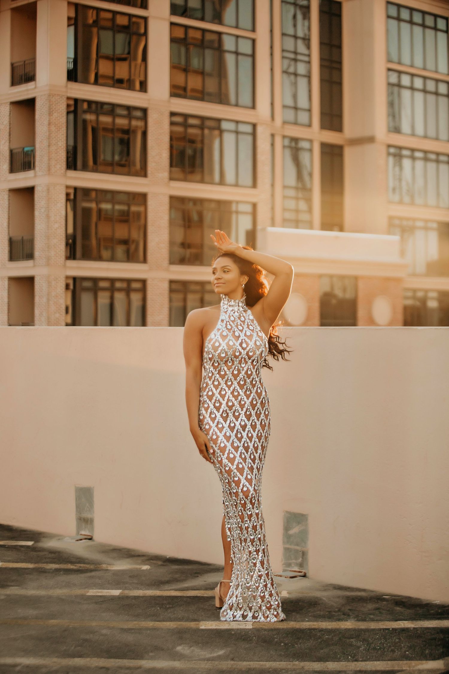 Person in sparkly formal gown poses gracefully against modern building at sunset.