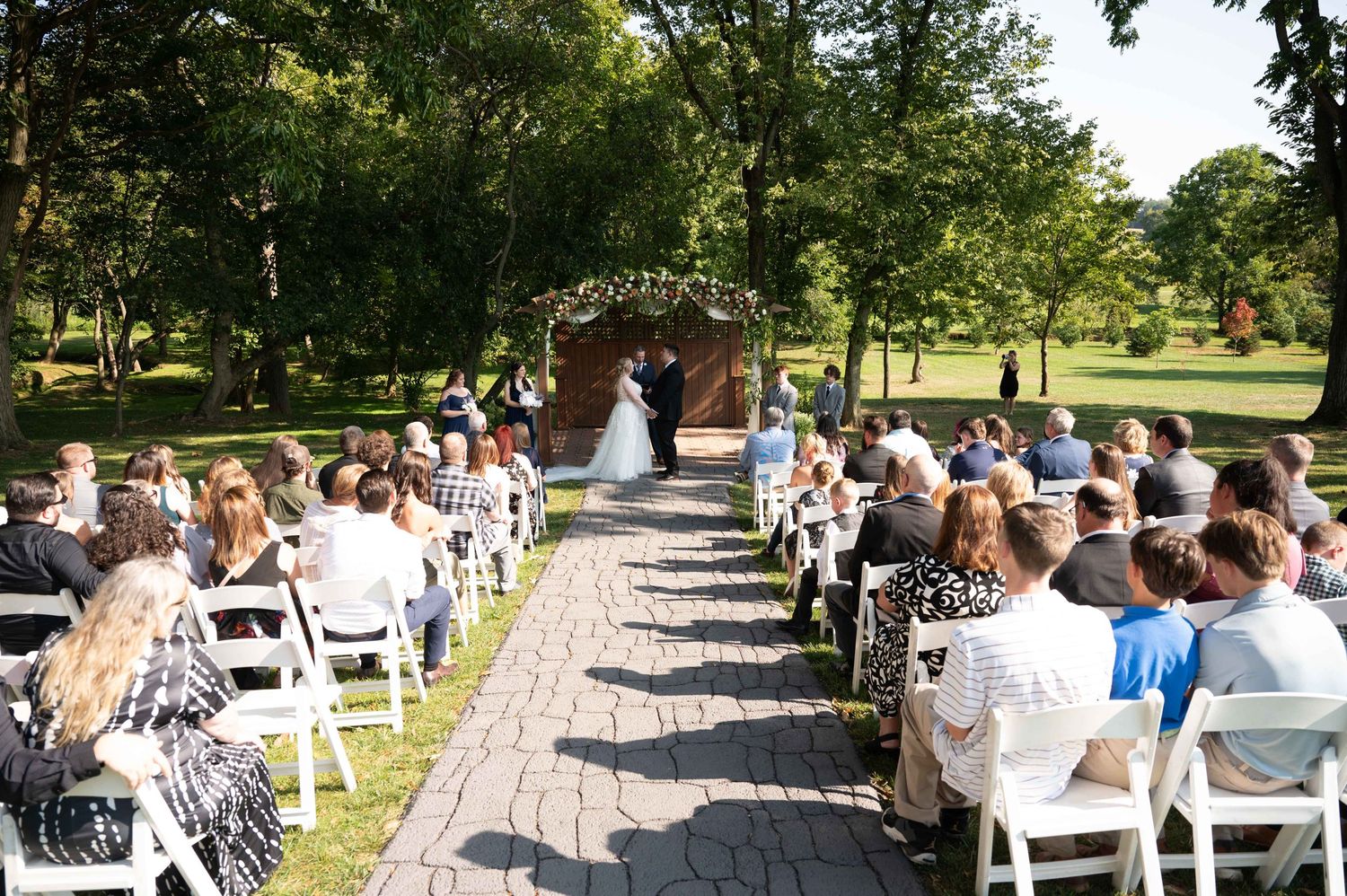 Outdoor wedding ceremony with guests seated on white chairs along brick pathway leading to floral arch.