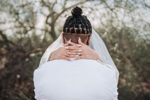 Close-up photo of hands touching a neck with braided hair and wedding veil against a blurred outdoor background.