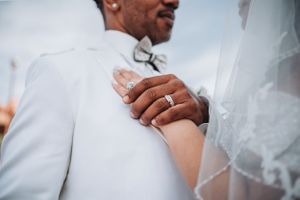 Close-up of wedding rings and hands against white formal attire.