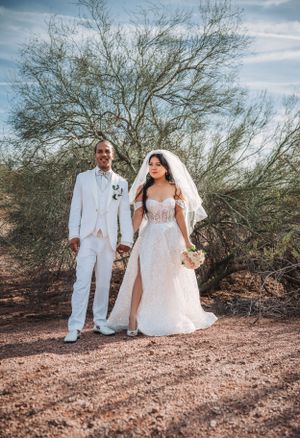 Wedding couple in all white attire pose together in desert landscape with barren trees.