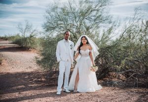 Newlyweds in elegant white outfits pose together on desert trail with sparse vegetation.