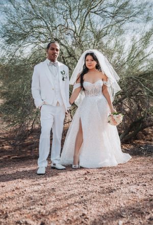 Wedding couple in white attire standing together on desert path with desert flora background.