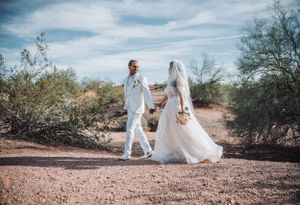Couple in white wedding attire walking together on desert trail under blue sky.