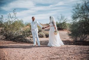Wedding couple in white attire poses on desert path against cloudy sky and desert brush landscape.
