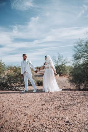 Desert wedding scene with couple in white standing on sandy path under blue sky.