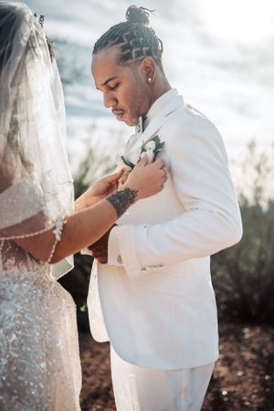 Groom in crisp white suit has boutonniere pinned during outdoor desert wedding ceremony.