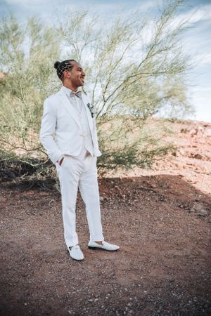 Groom in all-white suit stands confidently against desert landscape with wispy trees.