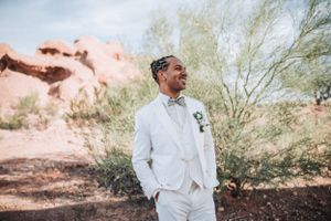 Desert wedding portrait of groom in white suit smiling against red rock formations.