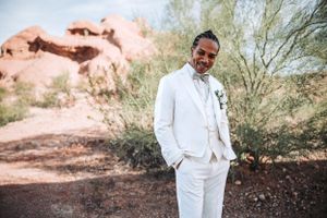 A groom in an all-white suit stands against a desert landscape with red rocks and desert vegetation in the background.