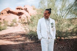 A stylish wedding portrait features a crisp white suit against the red rocks and desert brush of a natural landscape.