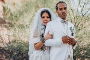 A wedding couple poses in white attire against desert scenery, with the bride wearing a sparkling gown and veil.