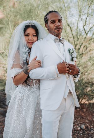 Newlyweds embrace in formal wedding attire against a desert backdrop with red rocks and desert plants.