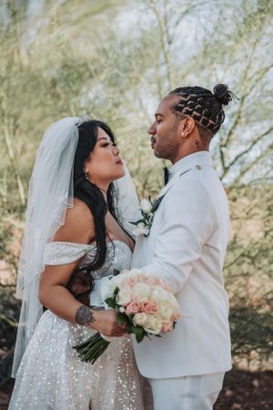 Elegant wedding portrait showing couple in white with peach bouquet against natural backdrop.