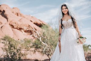 Bride in sparkly white gown poses against stunning red rock formations in desert landscape.