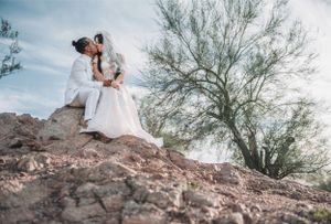 Newlyweds share romantic moment while perched on desert rocks under blue sky.