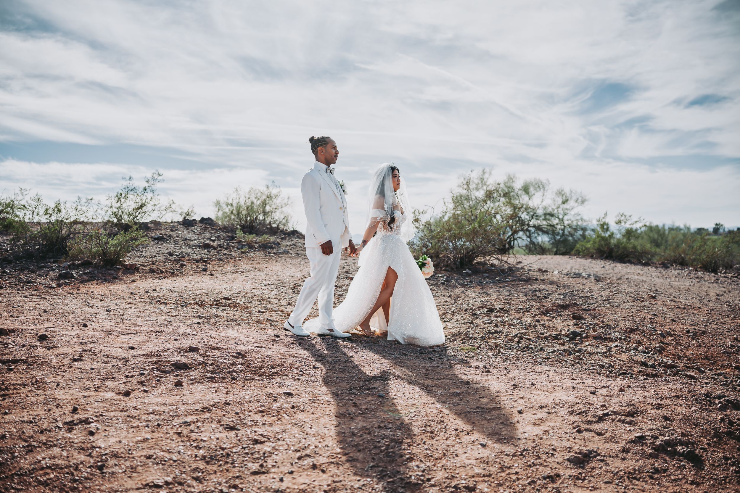 A wedding couple walks through a desert landscape under cloudy skies.