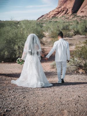 A wedding couple hold hands while walking on a desert trail with red rocks in the background.