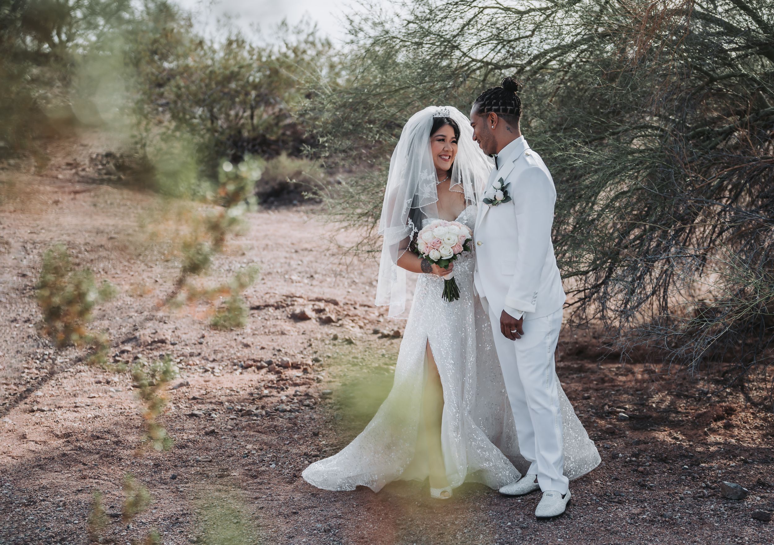A wedding couple in white outfits share an intimate moment on a desert path.