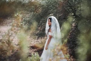 A bride in a white dress and veil poses among desert foliage.
