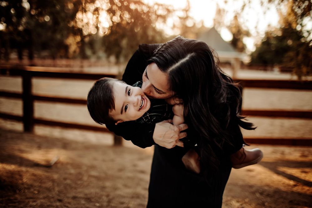A mother kisses her toddler son on a farm, standing near a rustic wooden fence, with a warm and peaceful countryside setting behind them.