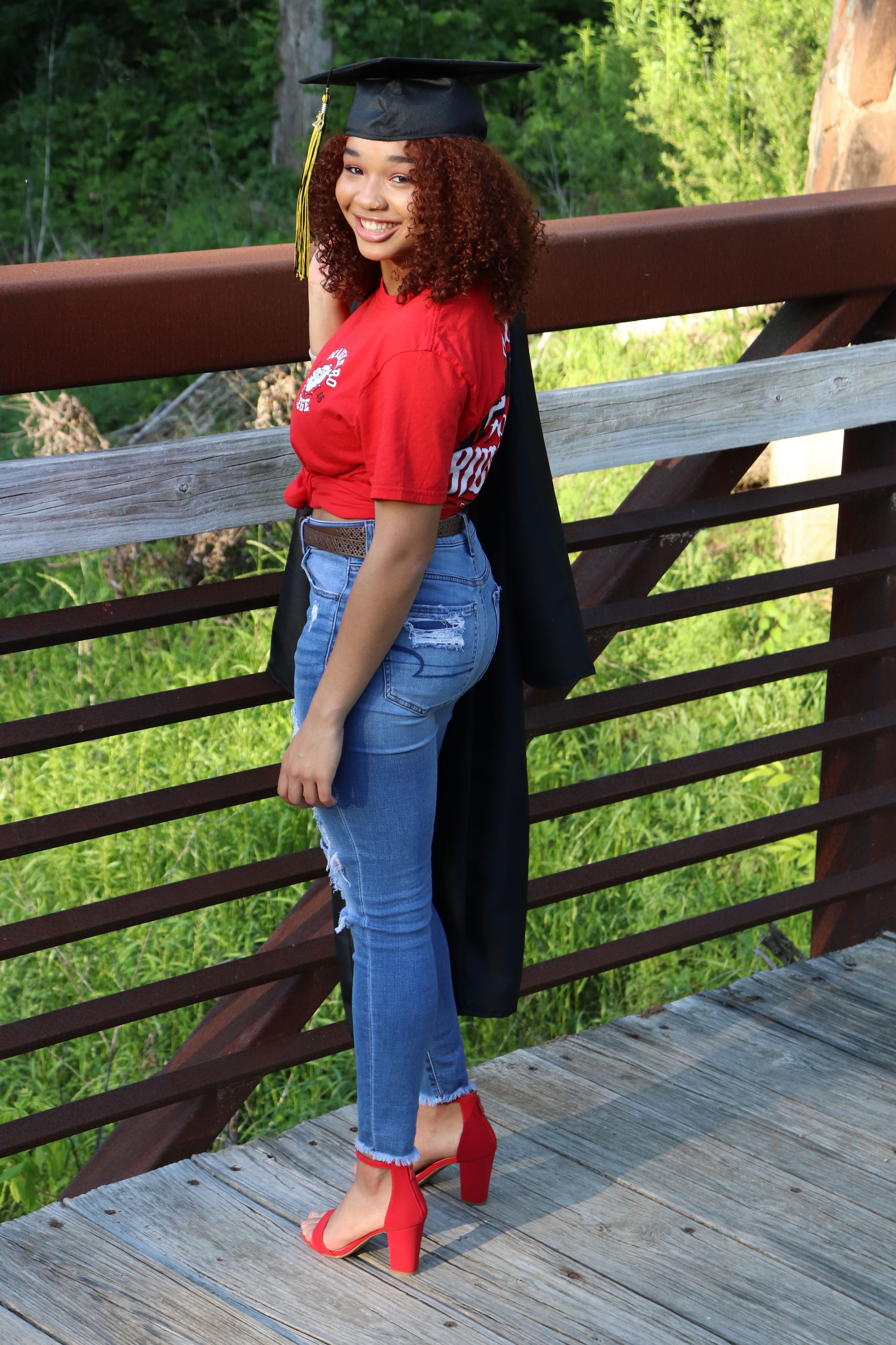 Graduate in red shirt and jeans poses on wooden bridge wearing academic regalia.