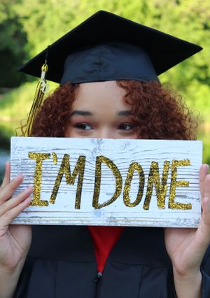 Graduate holds wooden sign reading "I'M DONE" while wearing cap and gown.