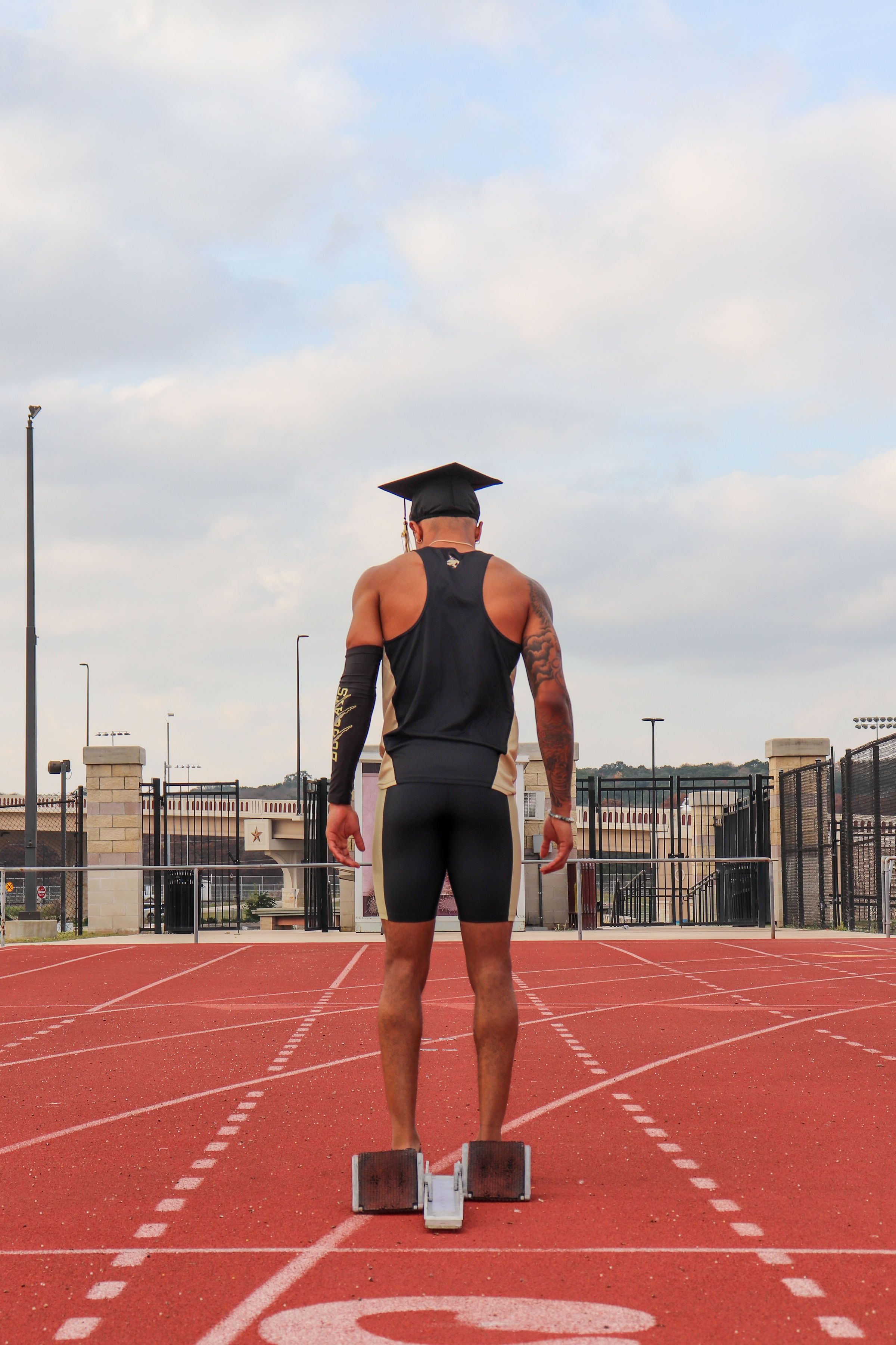 Graduate in athletic wear stands ready at starting blocks on a running track wearing a graduation cap.