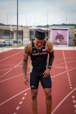 Athletic graduate in Texas State gear smiles while standing on a running track.