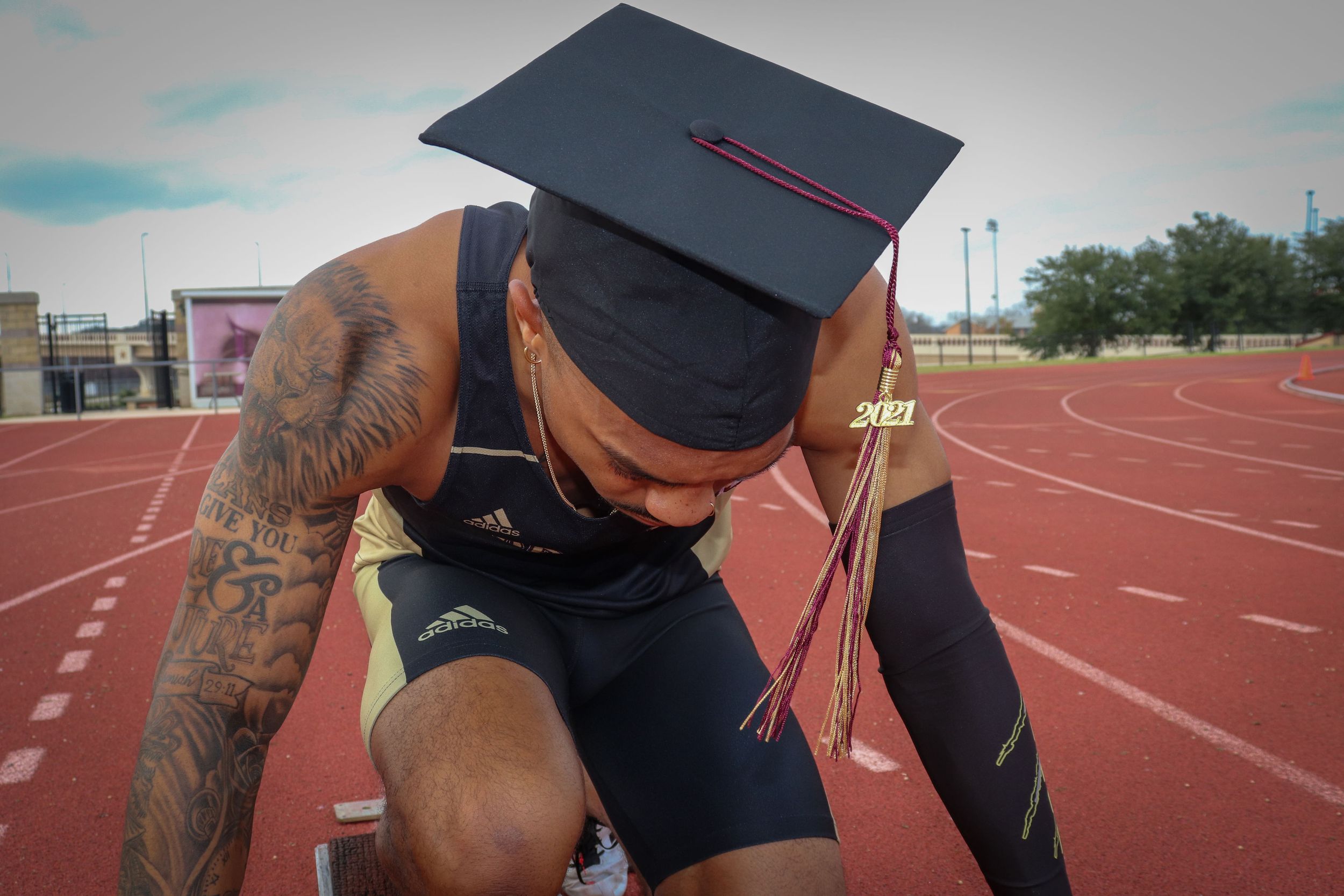 Graduate in athletic wear kneels on running track wearing cap and gown.