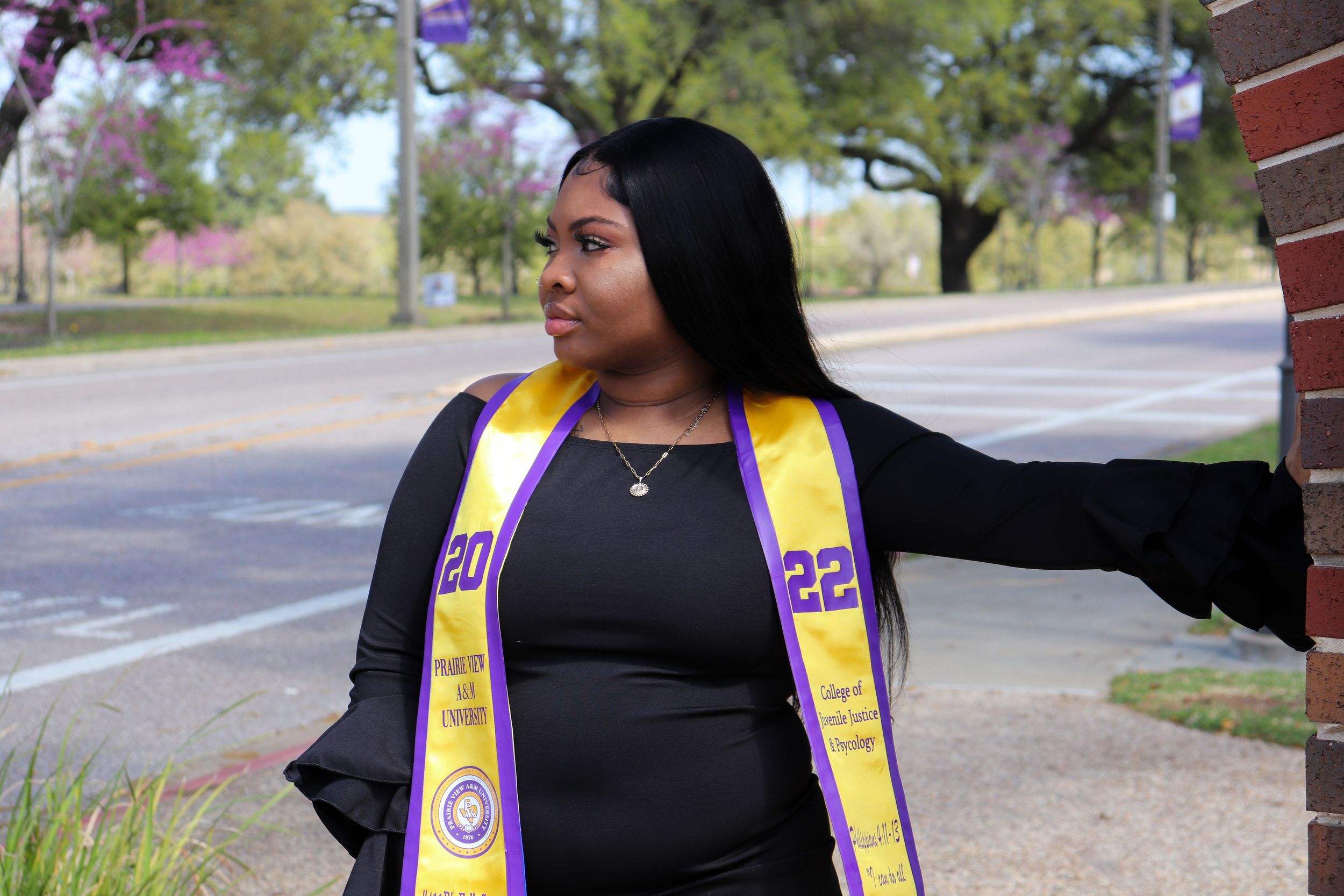 Graduate wearing academic stoles and black dress stands against brick wall outdoors.