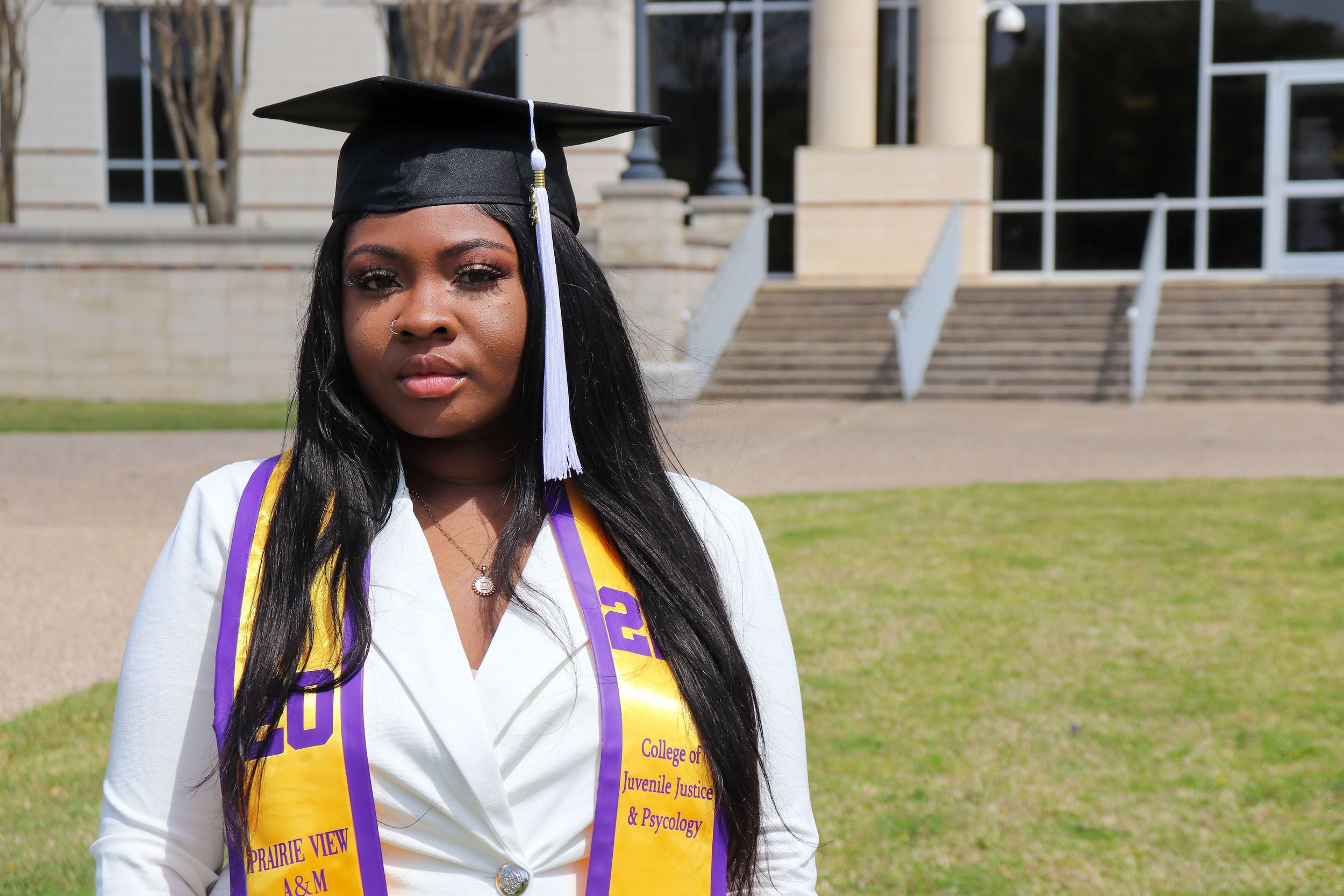 Graduate in cap and gown wearing yellow and purple honor cords poses outside academic building.