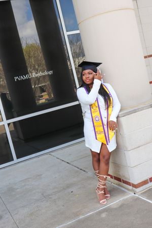 Graduate in white dress and academic regalia poses against university building column.