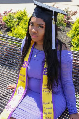 Graduate in purple dress and academic regalia with honor stole sits outdoors.