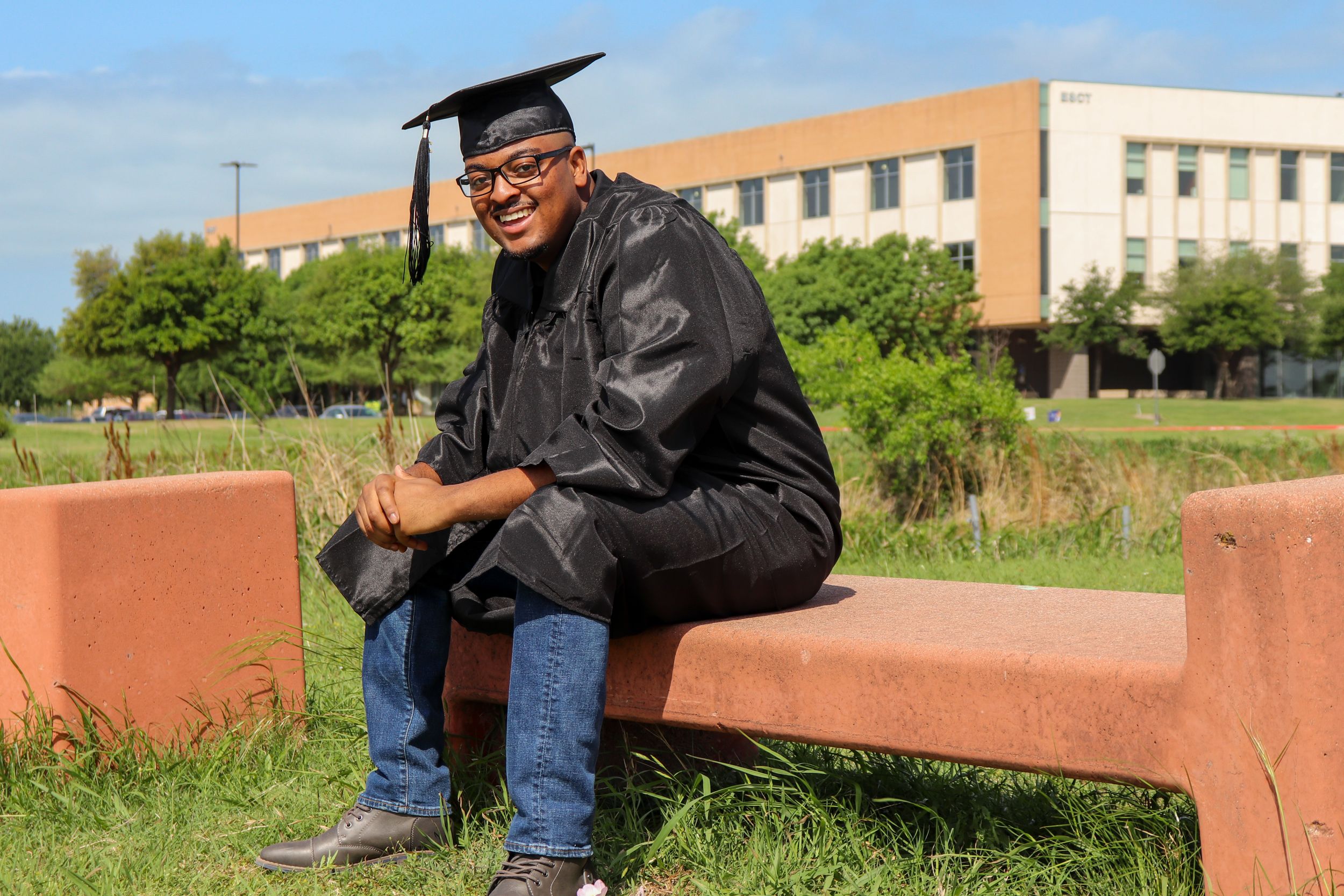 Graduate in traditional cap and gown sits casually on outdoor bench with campus building in background.