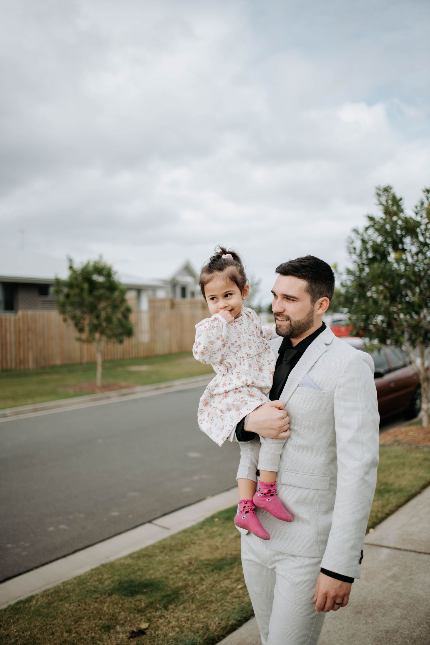 Member of wedding party in light suit holds small child wearing pink shoes on suburban street.