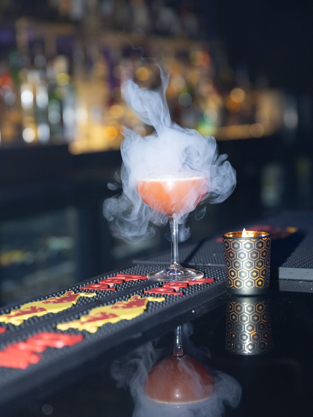 Dramatic smoky cocktail with orange glow on dark bar counter with decorative runner.