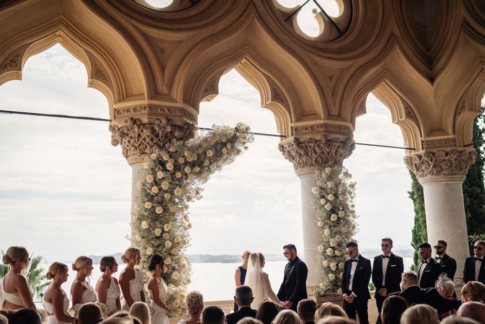 A wedding ceremony under Gothic arches decorated with white flowers overlooking a lake.