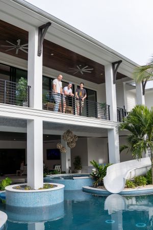 Three people standing on a luxury home balcony overlooking a stunning pool with a water slide.