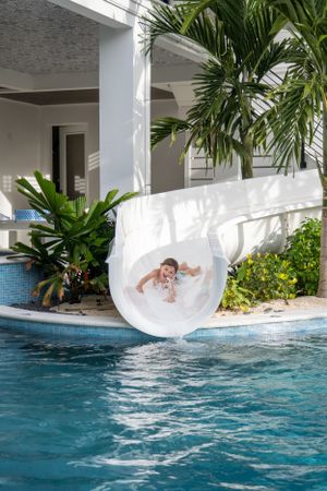 A smiling child emerges from a white water slide into a sparkling blue pool surrounded by tropical palms.