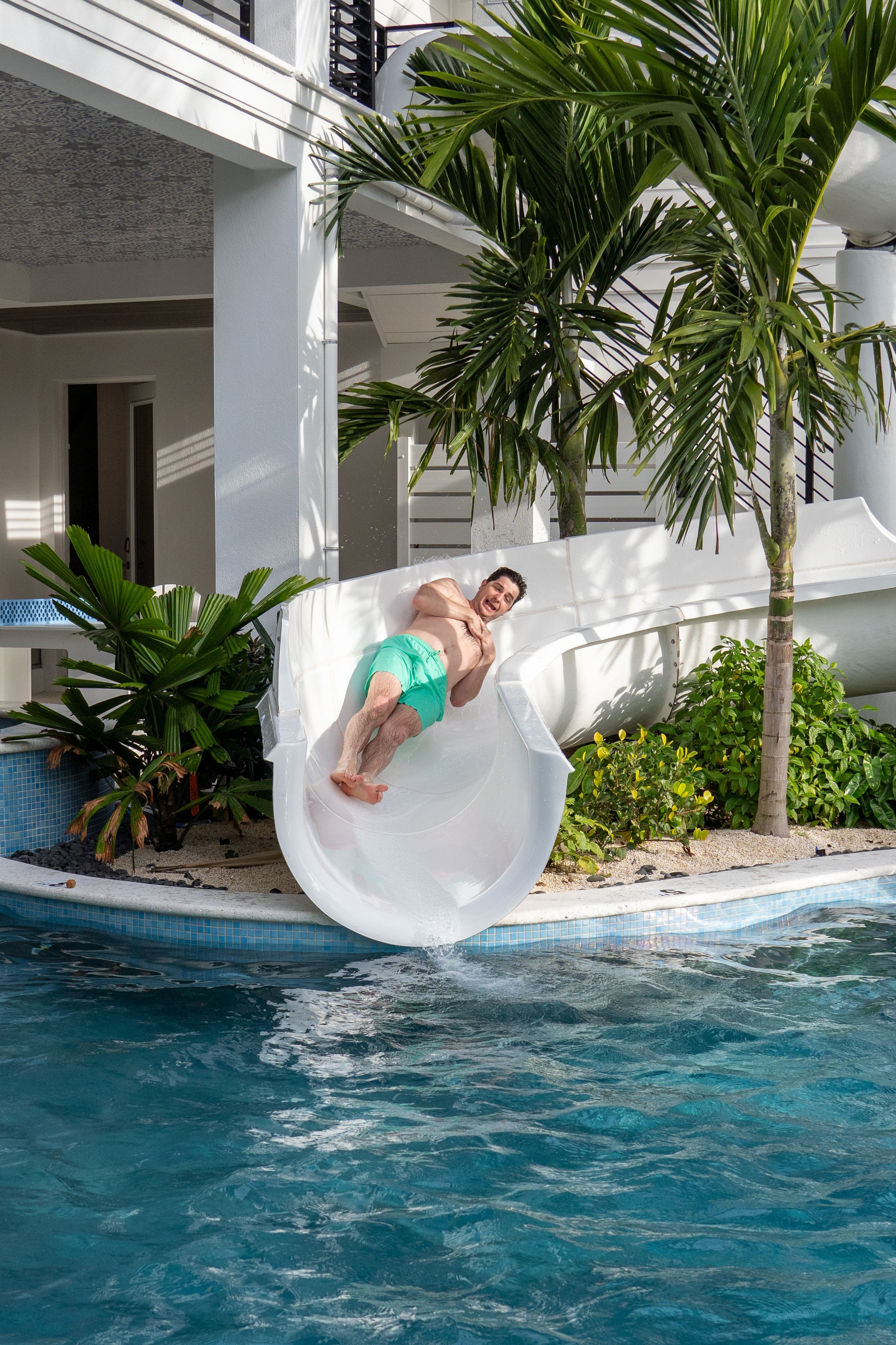 A man in turquoise swim trunks slides down a white water slide into a resort pool with tropical landscaping.