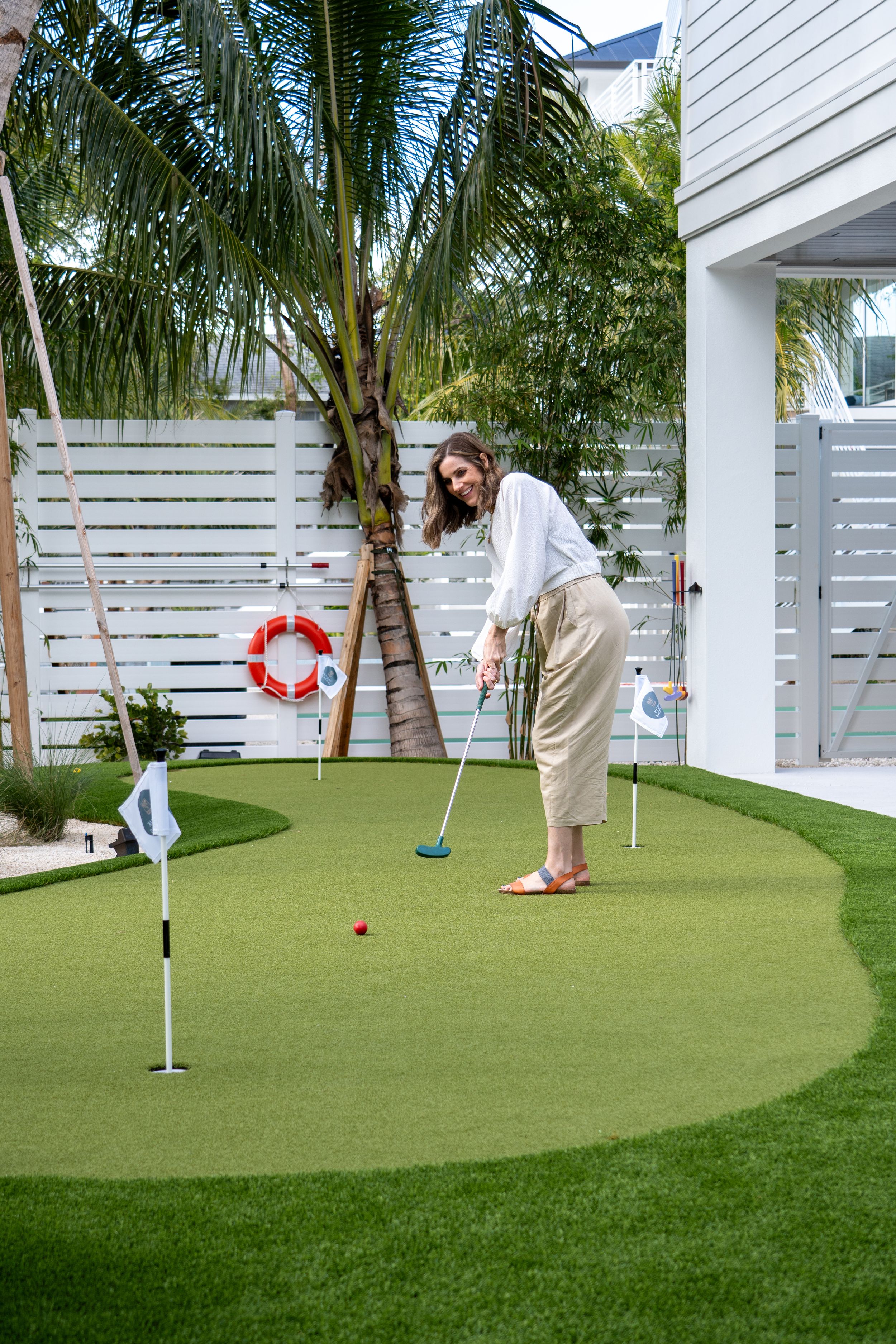 Woman in white top and beige pants playing mini golf on a backyard putting green with palm trees.