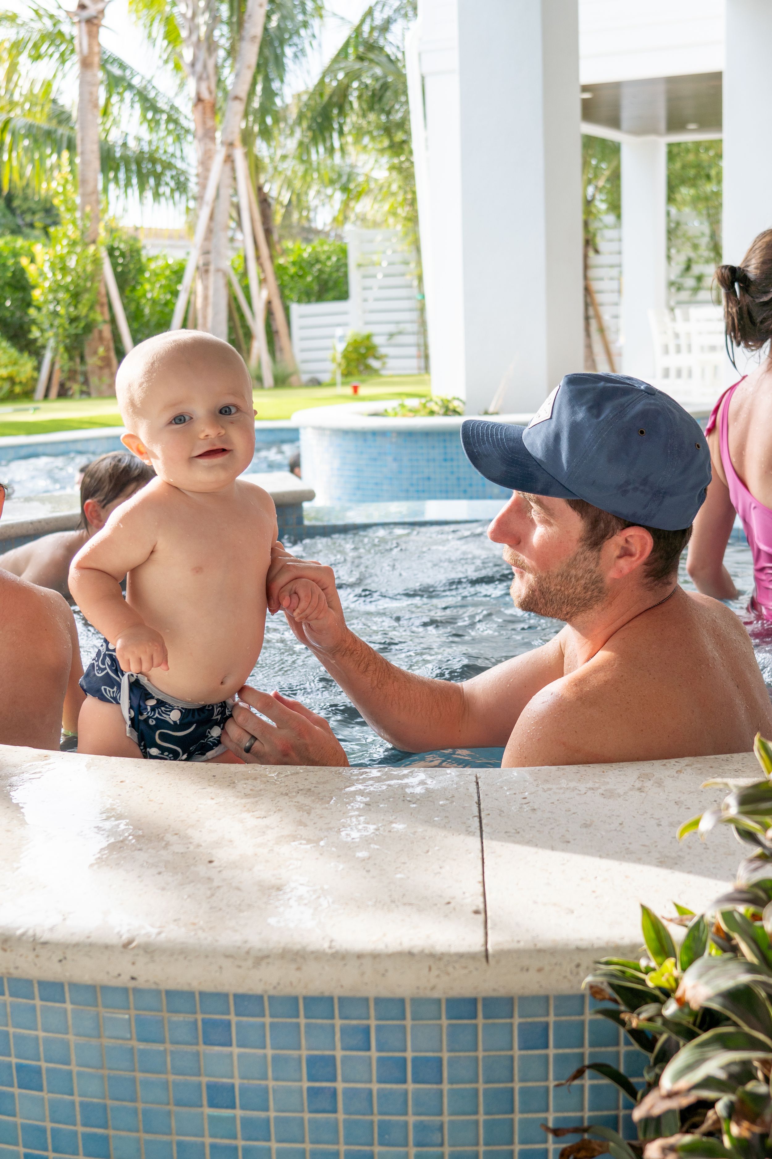 A smiling baby in swim trunks stands in a hot tub while a man in a blue cap holds him gently.