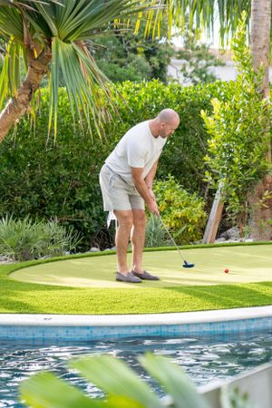A bald man practices putting on a small artificial green beside a resort pool surrounded by tropical plants.