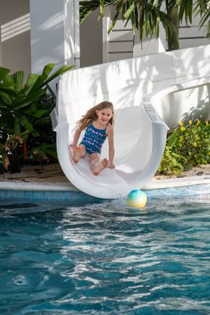 A laughing girl in a blue swimsuit slides down a white pool slide with a colorful ball in the water below.
