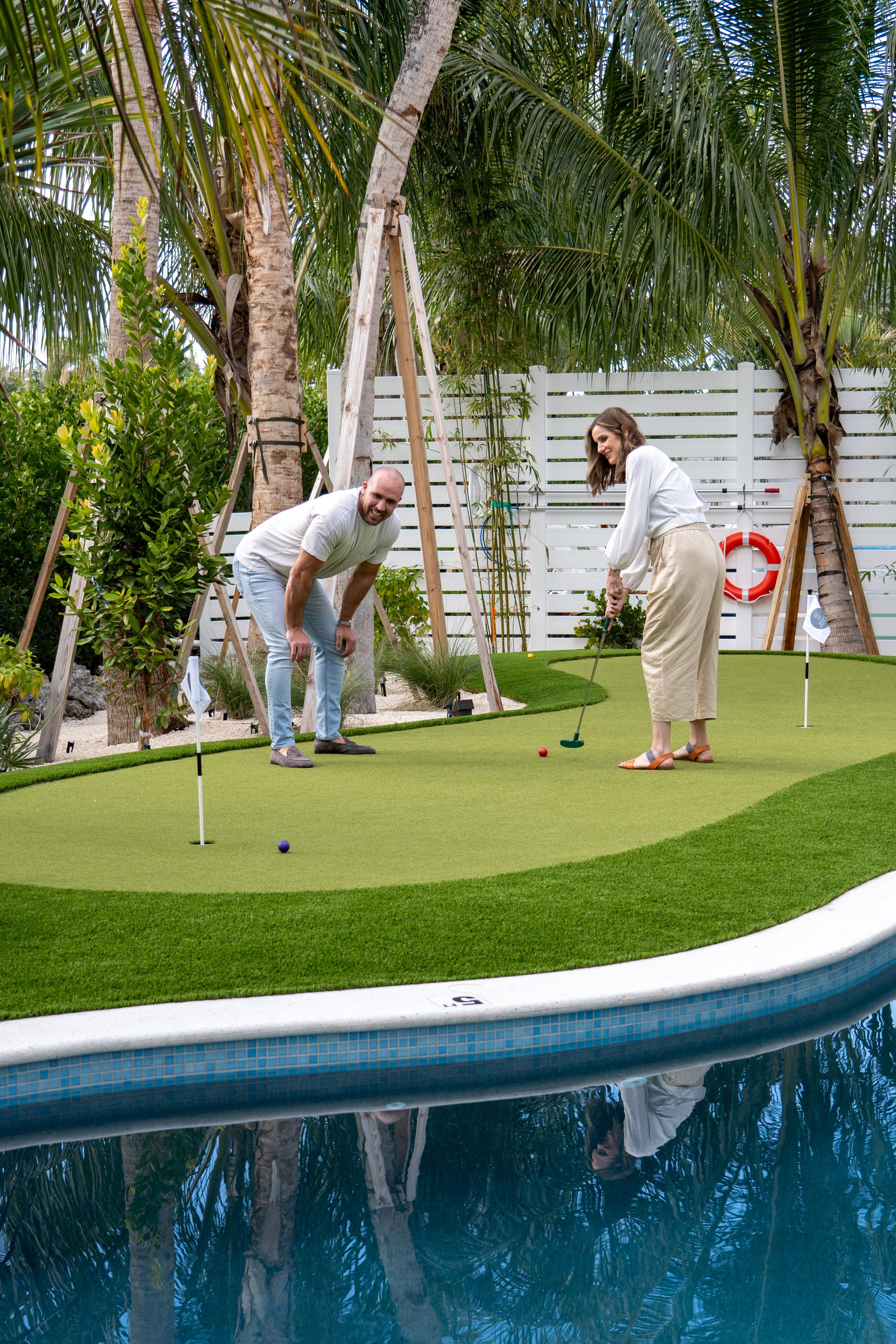 Couple playing mini golf on a private backyard putting green beside a pool with tropical landscaping.