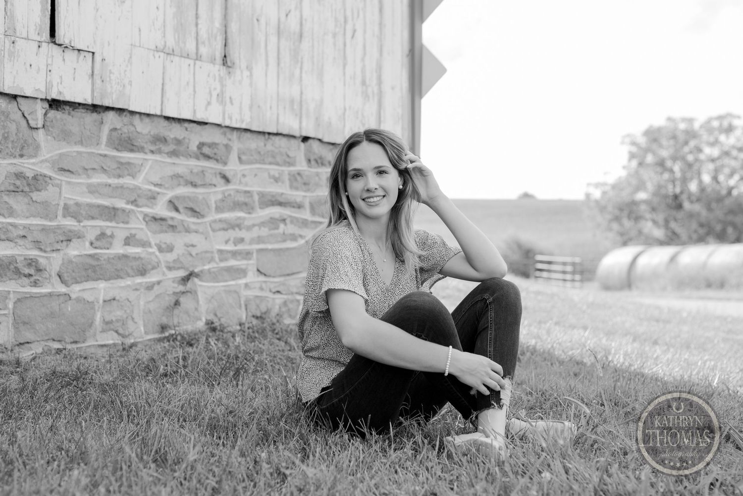 A black and white portrait of someone sitting by a stone wall in a pastoral landscape.